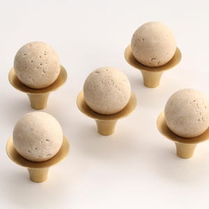 Könnte beinhalten: Fünf dekorative Schrankknöpfe. Jeder Knopf hat eine hellbeige, strukturierte Kugel auf einem goldfarbenen, ausgestellten Sockel. Die Knöpfe sind vor einem weißen Hintergrund angeordnet, um ihr Design hervorzuheben.