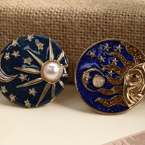May include: Two celestial-themed brooches. One features a sun with a pearl center and star accents on a dark blue background. The other has a crescent moon and stars with a sun face design. Both are gold-toned.