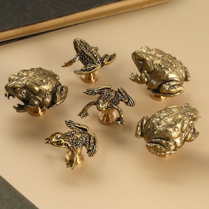Puede incluir: Seis tiradores de armario de latón antiguo con forma de rana en varias poses. Los tiradores están detallados con superficies texturizadas y están montados sobre pequeñas bases de latón. Las ranas están en diferentes posiciones.