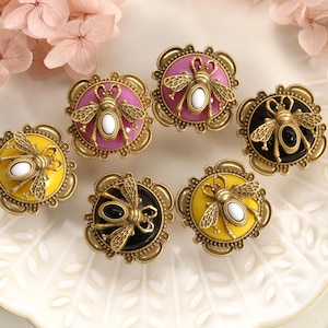 May include: Decorative cabinet knobs with a bee motif. The knobs feature a gold-tone metal frame with a circular design in yellow, pink, and black. Each knob has a bee in the center with a white or black oval. The knobs are arranged on a white, decorative plate.