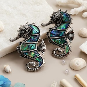 Peut inclure: Deux broches en forme d'hippocampe avec des incrustations nacrées vertes et bleues. Les broches ont une finition argentée et sont entourées de coquillages et d'éléments décoratifs, évoquant un thème côtier.