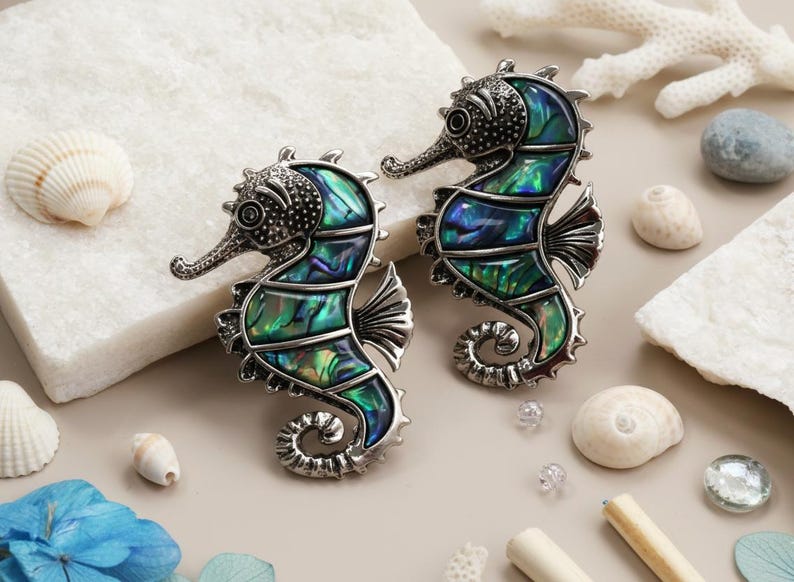 May include: Two seahorse brooches with iridescent green and blue abalone shell inlays. The brooches have a silver finish and are surrounded by seashells and decorative elements, evoking a coastal theme.