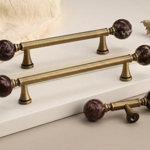 May include: Decorative cabinet pulls with a bronze finish and dark red marble-like sphere accents. The pulls are shown in a set of three, with two installed and one separate. These are ideal for adding a touch of elegance to furniture.