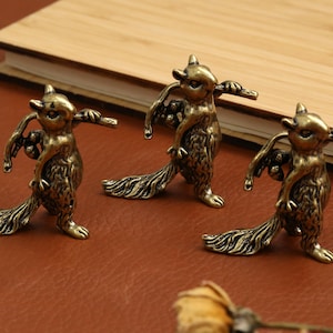 Könnte beinhalten: Drei bronzene Eichhörnchenfiguren, die jeweils einen Zweig mit Beeren halten. Die Eichhörnchen haben detailliertes Fell und lange, buschige Schwänze. Sie sind auf einer braunen Oberfläche mit einem hölzernen Notizbuch im Hintergrund platziert.