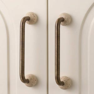 Könnte beinhalten: Zwei Schrankgriffe mit Bronze-Finish und einer steinartigen Kugel an jedem Ende. Die Griffe sind gebogen und an einer weißen Schranktür befestigt. Die Steinkugeln haben ein gesprenkeltes Aussehen.