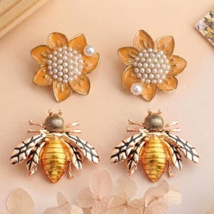 May include: Four decorative earrings: two sunflower-shaped and two bee-shaped. The sunflower earrings are orange with a pearl centre and a single pearl accent. The bee earrings are gold and black with detailed wings and bodies.