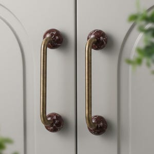 Könnte beinhalten: Zwei Schrankgriffe mit Messing-Finish und dunkelroten, marmorähnlichen Kugeln an jedem Ende. Die Griffe sind an einer hellgrauen Schranktür befestigt. Das Design ist eine moderne Interpretation traditioneller Hardware.