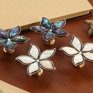 May include: Four flower-shaped cabinet knobs. Two knobs have iridescent blue and green petals, and two have white petals. Each knob has a silver trim and a gold base. The knobs are arranged on a brown surface.