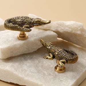 May include: Two brass alligator-shaped cabinet knobs. The knobs have detailed scales and are mounted on a round base. They are displayed on white stone blocks against a neutral background.