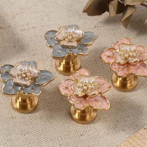 May include: Four flower-shaped cabinet knobs with gold bases. The flower designs feature petals in shades of pink and blue, with a cluster of faux pearls at the centre. These knobs are suitable for adding a touch of elegance to furniture or decor.