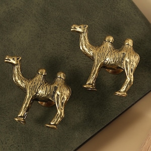 Puede incluir: Dos tiradores de armario antiguos de latón con forma de camello. Los tiradores tienen un acabado texturizado y están montados sobre una superficie verde oscuro. Los camellos están orientados en direcciones opuestas.