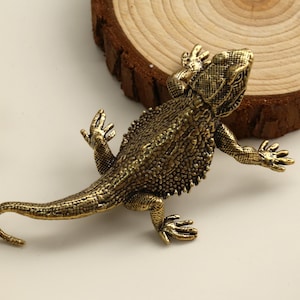 Puede incluir: Una detallada figura de lagarto de latón de estilo antiguo con piel texturizada y extremidades extendidas. El reptil está posicionado sobre una superficie blanca, cerca de una rodaja de madera, mostrando su intrincado diseño y tono dorado.
