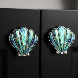 May include: Two seashell-shaped cabinet knobs with iridescent green, blue, and silver accents. The knobs are mounted on a black cabinet door, offering a coastal or nautical theme.