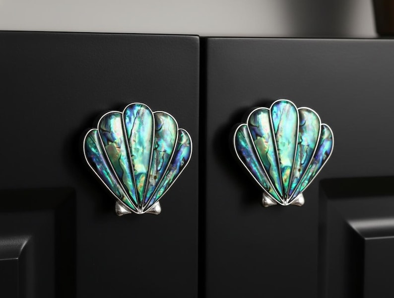 May include: Two seashell-shaped cabinet knobs with iridescent green, blue, and silver accents. The knobs are mounted on a black cabinet door, offering a coastal or nautical theme.