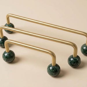 May include: Three cabinet pulls featuring a brushed gold metal handle with a U-shaped design. Each end of the handle is attached to a spherical dark green marble base with visible natural veining, creating a distinct contrast between the metal and stone.