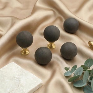 May include: A collection of five dark gray spherical cabinet knobs with brass bases. The knobs are arranged on a satin fabric, with a marble tile and eucalyptus sprig in the background. The knobs are a decorative hardware item.