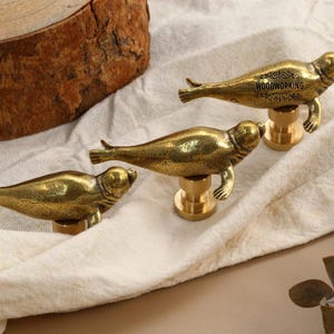 May include: Three brass seal-shaped cabinet knobs on a white cloth. The knobs are detailed with flipper and eye features. One knob has the word "WOODWORKING" engraved on it. The background includes a wood slice and a sprig of leaves.