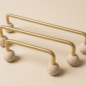 May include: Three gold-colored cabinet pulls with a curved bar design. Each pull is supported by a spherical, textured, beige-colored base. The pulls are arranged in a staggered, parallel configuration.