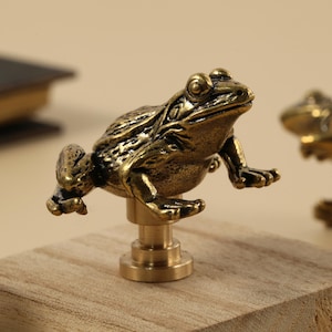 Op de afbeelding: Gedetailleerd messing kikkerbeeldje gemonteerd op een houten blok. De kikker zit met uitgestrekte voorpoten. Het beeldje heeft een vintage, antieke uitstraling.