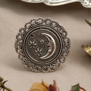 May include: Silver-toned metal drawer pulls with a celestial design. The central pull features a crescent moon and three stars within a circular frame, surrounded by an ornate, filigree-style border. Other hardware is visible in the background.