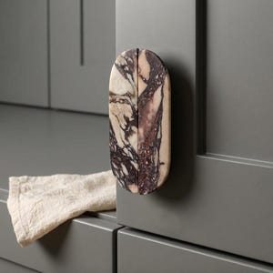May include: An oval-shaped cabinet knob made of polished marble with a distinct veined pattern in shades of deep purple, dark brown, and cream. The knob is mounted vertically on a flat, matte grey cabinet door surface with a clean, smooth finish.