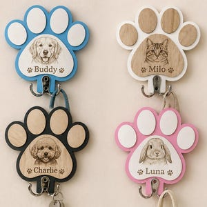 Puede incluir: Cuatro llaveros en forma de pata en azul, blanco, negro y rosa. Cada uno presenta un retrato de mascota y un nombre: Buddy, Milo, Charlie y Luna. Incluye ganchos para llaves. De madera.