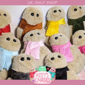 May include: A group of ten plush toys shaped like amoebas, each wearing a different coloured scarf. The scarves are red, brown, yellow, green, light blue, pink, black, and a sparkly pink. The toys are all beige and have large black eyes. The text "Kitty's Creatives" is visible in the bottom centre of the image.