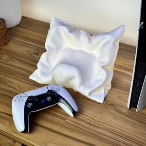 May include: A white and black game controller rests on a wooden surface next to a white, abstract-shaped decorative object. A white game console is visible in the background. The controller has black joysticks and buttons.