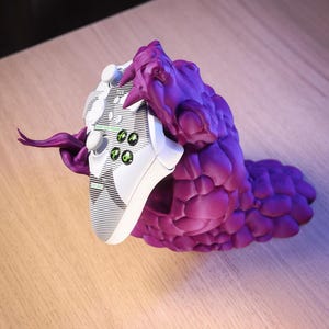 May include: A purple 3D printed monster holding a white video game controller. The controller has green buttons and a white faceplate with black lines.