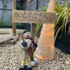 May include: Wooden sign with the text "SANTA, PLEASE STOP HERE FOR Rory, Eva & Idah" with a drawing of Santa in a sleigh pulled by reindeer. The sign is attached to a wooden post with a snowman figurine standing next to it.