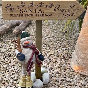 May include: Wooden sign with a Christmas theme, featuring a reindeer-drawn sleigh and the text "Santa, please stop here for Rory, Eva & Lilah". A snowman figurine stands in front of the sign.