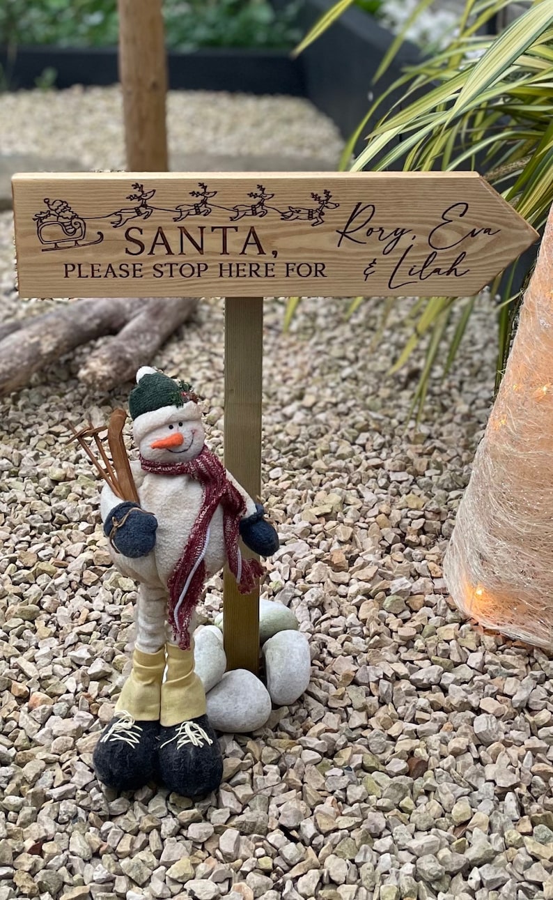 May include: Wooden sign with a Christmas theme, featuring a reindeer-drawn sleigh and the text "Santa, please stop here for Rory, Eva & Lilah". A snowman figurine stands in front of the sign.