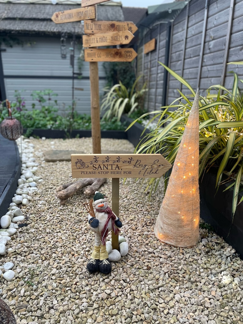May include: A wooden sign with the text "Please stop here for Santa" and a snowman figurine standing on a bed of gravel.