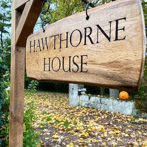 May include: A wooden sign with the words "HAWTHORNE HOUSE" carved into it. The sign is hanging from a wooden post with metal chains. The sign is in front of a brick wall with a small pumpkin sitting on top of it.