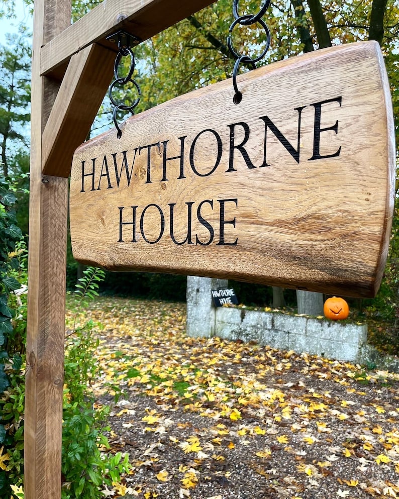 May include: A wooden sign with the words "HAWTHORNE HOUSE" carved into it. The sign is hanging from a wooden post with metal chains. The sign is in front of a brick wall with a small pumpkin sitting on top of it.