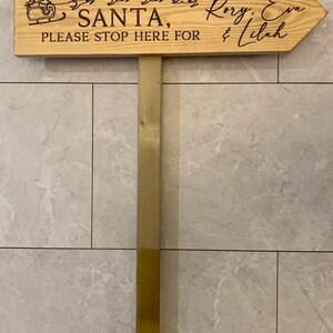 May include: Wooden sign with a Santa Claus and reindeer design. The sign reads "Santa, please stop here for Rory, Eva & Lilah".
