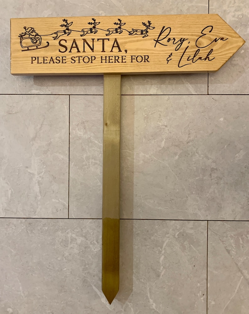 May include: Wooden sign with a Santa Claus and reindeer design. The sign reads "Santa, please stop here for Rory, Eva & Lilah".