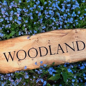 May include: A wooden sign with the word "WOODLAND" engraved in black lettering. The sign is lying on a bed of blue forget-me-not flowers.