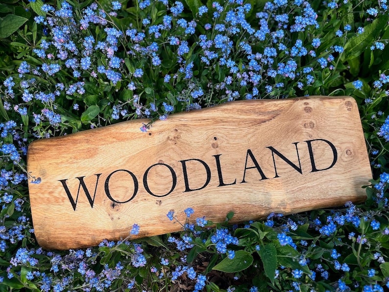 May include: A wooden sign with the word "WOODLAND" engraved in black lettering. The sign is lying on a bed of blue forget-me-not flowers.
