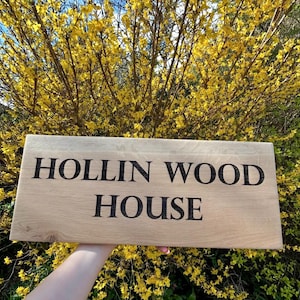 May include: A wooden sign with the text "HOLLIN WOOD HOUSE" engraved in black lettering. The sign is held in front of a bush with yellow flowers.