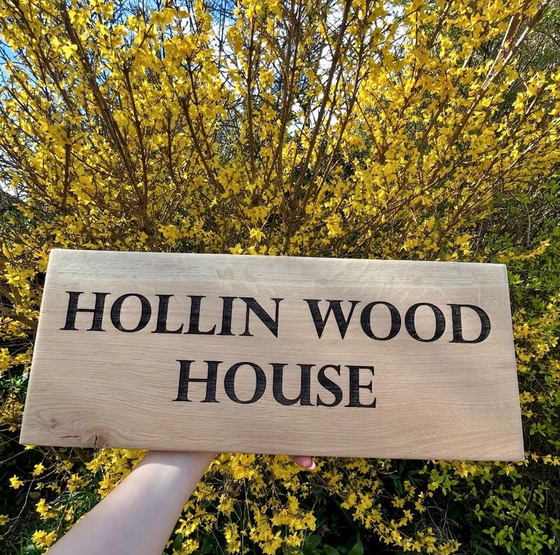 May include: A wooden sign with the text "HOLLIN WOOD HOUSE" engraved in black lettering. The sign is held in front of a bush with yellow flowers.