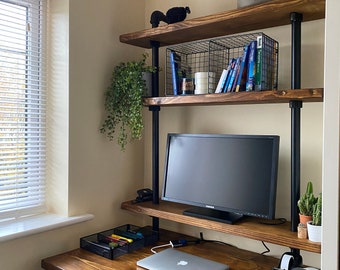 May include: A wooden desk with black metal legs and a shelf unit above. The desk has a laptop, a monitor, and a black wire basket with colorful markers. The shelf unit has a black wire basket, books, and a potted plant.