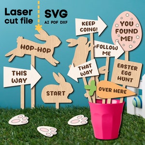Puede incluir: Señales de madera con temática de Pascua con flechas y recortes de conejos, incluyendo "Hop-Hop", "This Way" y "Easter Egg Hunt". Una taza rosa sostiene las señales, colocadas sobre hierba verde con una valla blanca y huevos de Pascua en el fondo. El texto "Laser cut file SVG" está en la parte superior.