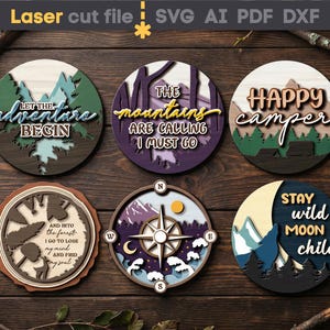 Puede incluir: Un conjunto de seis posavasos de madera cortados con láser con diseños temáticos de la naturaleza. Cada posavasos presenta un diseño único con frases como "Let the adventure begin" y "Happy camper". Los posavasos están dispuestos sobre una superficie de madera oscura.