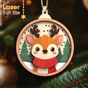 May include: A circular wooden Christmas ornament featuring a layered design of a cartoon deer with brown antlers, pink ears, and a red scarf. The background displays green pine trees and white snow against a light wood frame with text Laser cut file.