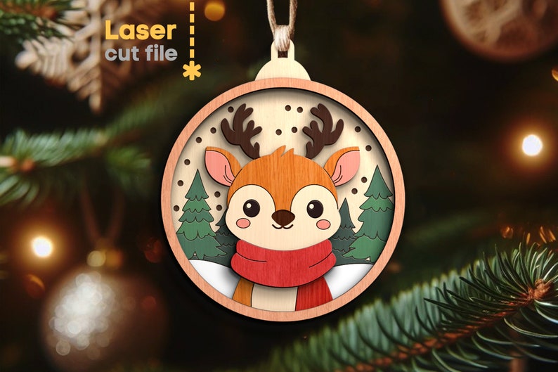 May include: A circular wooden Christmas ornament featuring a layered design of a cartoon deer with brown antlers, pink ears, and a red scarf. The background displays green pine trees and white snow against a light wood frame with text Laser cut file.