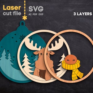 May include: A three-layer laser cut file for a Christmas ornament featuring a teal background with pine trees and snowflakes, a light wood layer with a moose, and a wood ring layer with a brown moose nose and a yellow scarf. Text reads Laser cut file SVG AI PDF DXF 3 LAYERS.