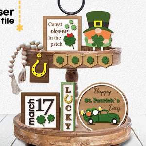 May include: A tiered wooden tray decorated with St. Patrick's Day themed laser cut wood signs. The signs feature a leprechaun, a horseshoe, a truck with shamrocks, a clover, and the text "Cutest clover in the patch", "March 17", "LOCKY", and "Happy St. Patrick's Day".