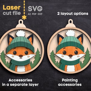 May include: A laser cut file SVG featuring two circular wooden ornament designs of a fox wearing a green knit hat with a white pom-pom and a matching green scarf. The fox is set against a background of green pine trees. Text includes Laser cut file, SVG, AI PDF DXF, 2 layout options, Accessories in a separate layer, and Painting accessories.