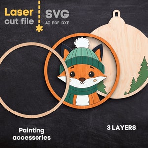 May include: A laser cut file SVG design for a three-layer wooden ornament featuring a fox wearing a teal beanie and scarf. The design includes a circular frame, a fox cutout, and a back piece with pine tree silhouettes. Text reads Laser cut file, SVG, AI, PDF, DXF, 3 LAYERS, and Painting accessories.